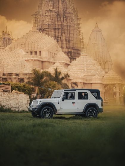 A side profile of the white Mahindra Thar on a lush green field, with the temple architecture in the background. This composition highlights the car's strong presence and versatility, equally at home in both rugged and serene environments.