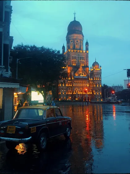 A classic Kaali-Peeli taxi parked on a flooded street at night, with the lights of the BMC building reflected in the water. This is a highly atmospheric and quintessentially Mumbai monsoon image.