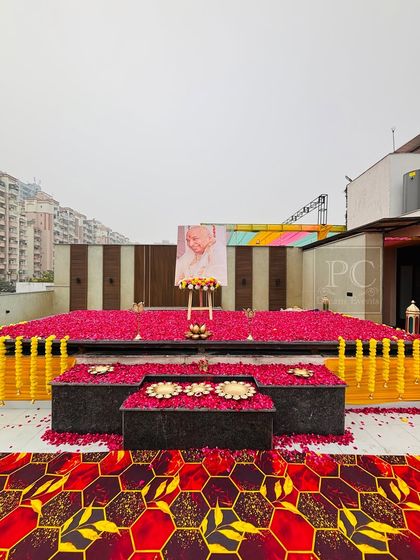 A wide view of the terrace satsang decor. The combination of the red petal-covered stage, marigold garlands, and geometric-patterned carpet creates a grand and devotional setting.