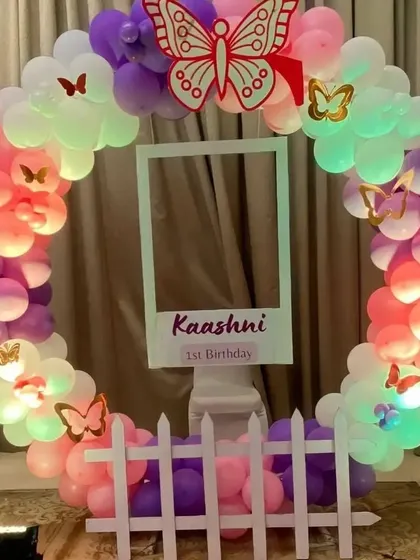 A butterfly-themed first birthday photo booth with a circular balloon frame and a polaroid-style cutout.