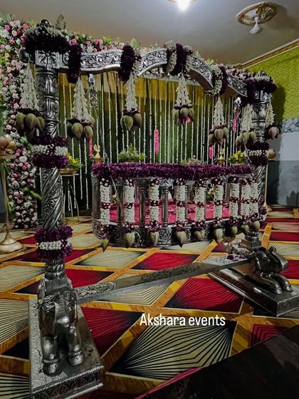 A grand silver cradle, intricately decorated with purple and white flowers, ready for a naming ceremony. The backdrop features hanging jasmine garlands for a classic touch.