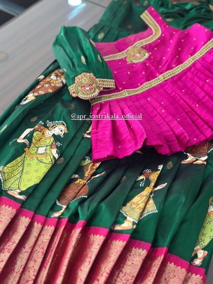 A unique hand-painted half-saree for kids. This dark green skirt features Kalamkari-style figures, paired with a vibrant pink pleated blouse for a striking contrast.