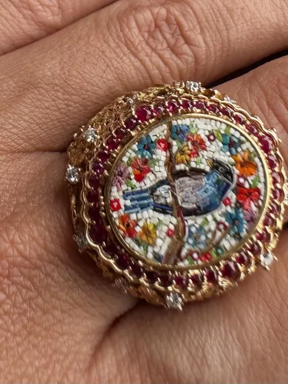 A close-up of a handcrafted ring featuring a bluebird among blossoms. The micromosaic is surrounded by a halo of deep red rubies and diamonds, set in an ornate 18k gold band.
