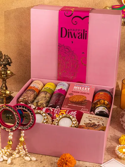 A pretty-in-pink Diwali hamper filled with savory and sweet treats. The warm candlelight and traditional brass lamp add a beautiful, ambient glow to the entire scene.