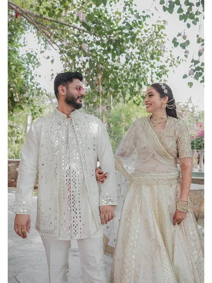 A candid walking shot of the couple sharing a happy glance. Dressed in coordinated white and silver mirror-work outfits, they look effortlessly elegant against the lush green backdrop of the resort.