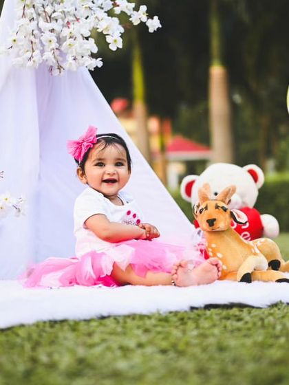 A cheerful outdoor picnic setup with a pink theme. It features a white teepee, pink and yellow balloons, plush toys, and lanterns, creating a soft and pretty scene on the grass.