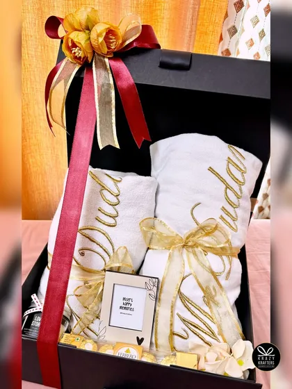 A luxurious wedding hamper for the new 'Mr. & Mrs.' featuring personalized towels with their names embroidered in gold, presented in a handsome black gift box with a red and gold ribbon.