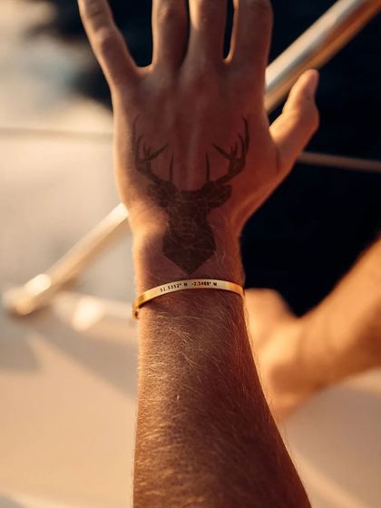 A man's arm with a prominent tattoo, complemented by a gold cuff bracelet engraved with coordinates. The bracelet adds a touch of personal, metallic detail to his style.