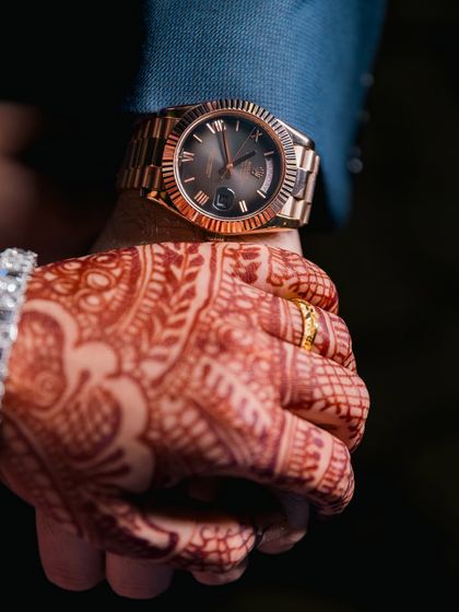 A close-up shot of the couple holding hands, highlighting the bride's mehendi and the groom's classic watch.