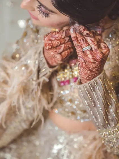 A detailed close-up of a bride adjusting her earring. The shot highlights her intricate henna design, her wedding ring, and the texture of her stunning sequined and feathered blouse.