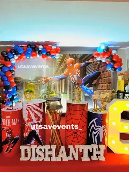 A Spiderman theme for Dishanth's 6th birthday. The setup includes a Spiderman backdrop, themed plinths, a red, blue, and black balloon arch, and a light-up number '6'.