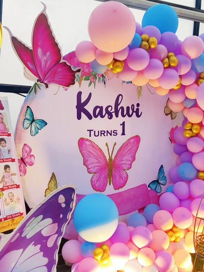 A close-up of a butterfly-themed first birthday, showing a photo collage board and a pastel balloon arch.