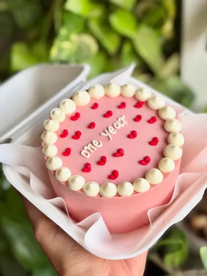 Tiny cake, big feels. This "one year" anniversary bento cake is dressed in a classic frosted pearl border and tiny red hearts.