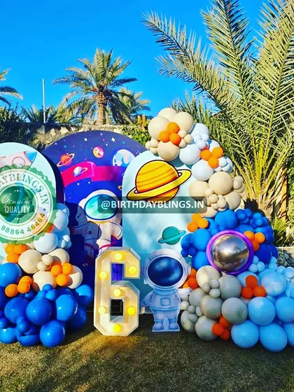 An outdoor space-themed party with a beautiful natural backdrop of palm trees. The setup includes multiple arched panels, astronaut cutouts, and a light-up number '6'.