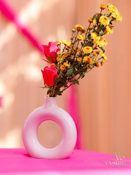 A simple, modern donut-shaped vase holding a single red rose and a sprig of yellow flowers, set on a bright pink surface.