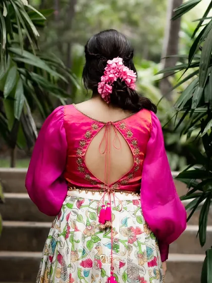 The back of this contemporary pink blouse features a keyhole opening and delicate embroidery, perfectly complementing the hand-painted Kalamkari lehenga for a modern yet traditional look.