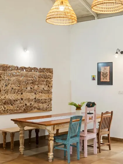A rustic dining area at Z Farms, featuring a long wooden table with mismatched colorful chairs. The carved wooden wall art and woven lampshades add to the eclectic, farmhouse charm.