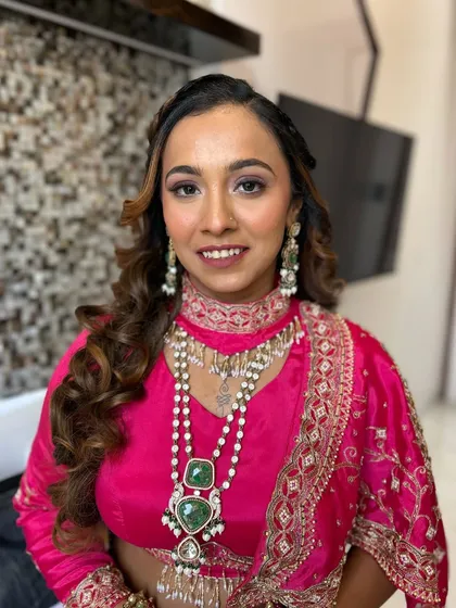 This client is radiant in her bright pink lehenga. The makeup features a soft smokey eye and a flawless base, complemented by a voluminous curly hairstyle.