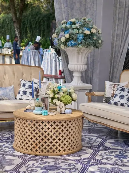 A lounge area at the French carnival, with classic beige sofas, cushions with palm tree prints, and a central wooden table with a floral centerpiece.