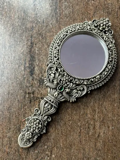 A beautifully crafted antique-finish hand mirror in 925 sterling silver. The detailed floral and peacock motifs, accented with a green stone, make this a timeless piece of art for your dressing table.