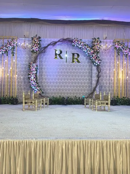 For this engagement, we designed a modern stage with a large central ring featuring the couple's initials. The backdrop includes gold-toned geometric panels and soft floral arrangements, all illuminated by warm bulb lighting.
