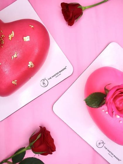 Two heart-shaped cakes, one in red with gold flakes and the other in pink with sugar pearls, both adorned with fresh roses. A perfect pair to celebrate love.