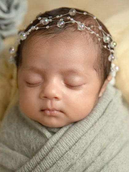 A close-up on the baby's face, showing their delicate features and the elegant crystal headband. The soft textures of the wrap and background add to the peaceful feel.