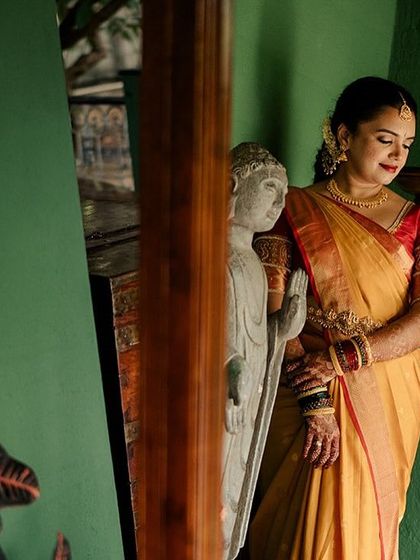A bride poses by a window, her reflection captured in the glass, next to a serene Buddha statue.