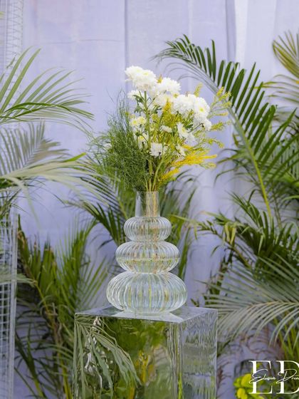 A minimalist floral arrangement in a modern, textured glass vase set against a backdrop of palm leaves. This simple yet elegant detail complements the larger botanical theme.