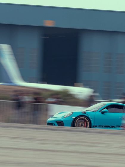 The same light blue Porsche, a masterpiece of German engineering, showing its track-focused aerodynamics as it speeds past the hangars.