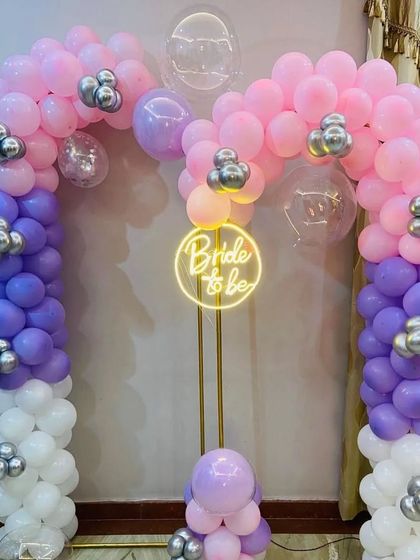 Here is another color option for a bride-to-be balloon arch. This setup uses a mix of pink, purple, and white balloons with silver accents, centered around a "Bride to be" neon sign on a stand for a softer, colorful look.