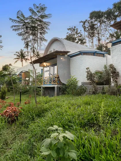 An exterior view of a Santorini-inspired villa in Coorg, with its whitewashed, curved walls nestled in a lush green landscape. The architecture offers a unique Mediterranean escape in the misty mountains.