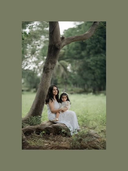A mother holds her child while sitting peacefully at the base of a large tree. The natural setting provides a serene backdrop for this intimate portrait, showcasing a quiet moment of connection.