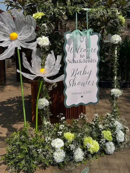 A personalized welcome sign for the baby shower, framed with lush greenery, white flowers, and whimsical oversized daisies to greet guests.