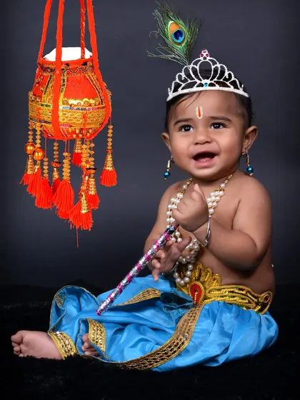 A joyful baby Krishna portrait against a simple dark background. The focus here is on the cute expression and the detailed costume, including the crown and pearl jewelry.