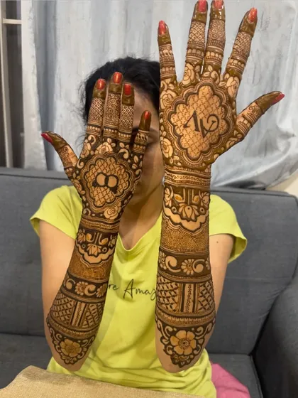 A happy bride showing her full-arm mehndi. One hand features the couple's initials 'AV', while the other has a symbol of togetherness, all tied together with floral bands.