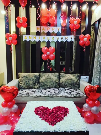 An elegant anniversary decoration in a living room space. The setup features a "Happy Anniversary" banner, red balloon arrangements, and a large heart made from rose petals on the center table.