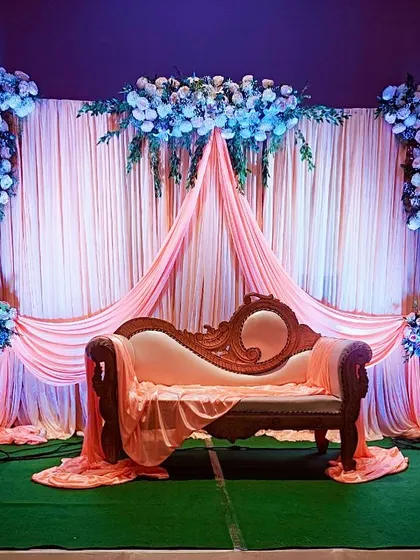 An elegant engagement stage setup with soft peach draping against a darker wall, highlighted by beautiful white and blue floral arrangements and a classic carved sofa for the couple.