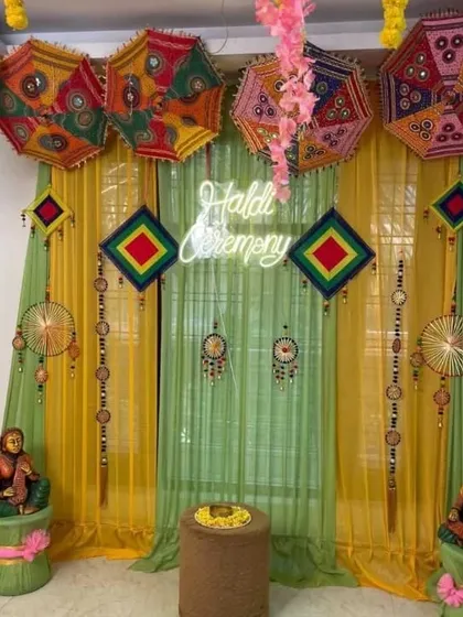 A detailed Haldi ceremony backdrop combining traditional and modern elements. The setup uses yellow and light green drapes, colorful Rajasthani umbrellas, and a central "Haldi Ceremony" neon sign to create a bright and energetic atmosphere.