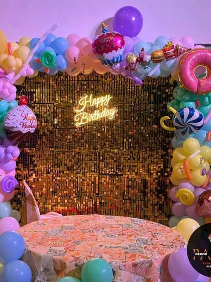 A candy-themed cake-cutting stage. The backdrop is a gold shimmer wall, framed by a balloon arrangement featuring various candy and donut-shaped foil balloons.