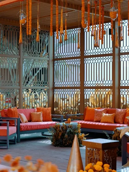 A comfortable lounge area with coral-colored sofas set against a backdrop of intricate jali screens and hanging marigold tassels.