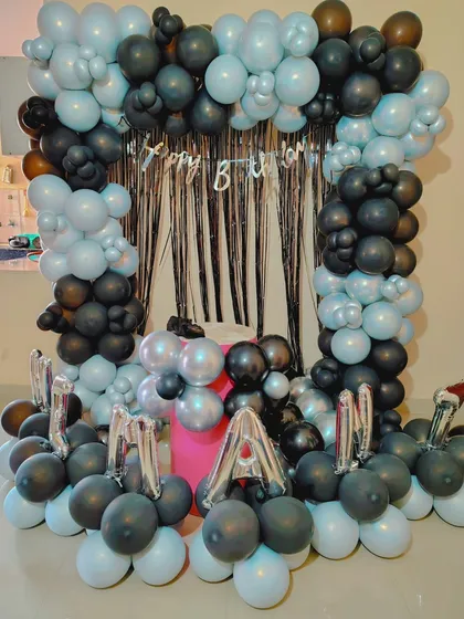 A birthday decoration using a frame of black and light blue balloons, with the name "MANAN" spelled out in foil letters.
