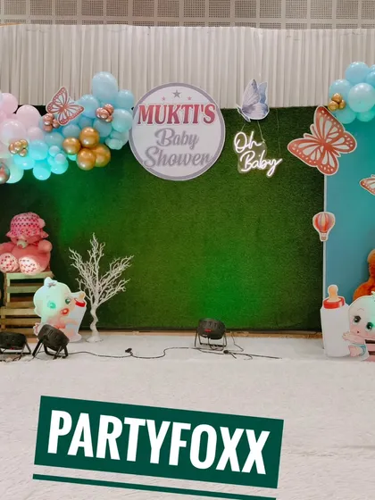 Mukti's baby shower featuring a green hedge wall, a pastel balloon arch, butterfly cutouts, and a neon "Oh Baby" sign.