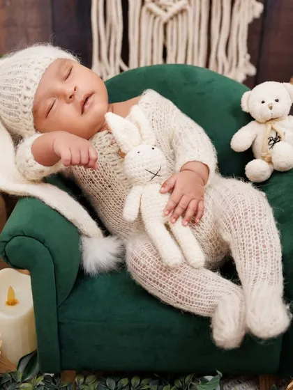 A newborn in a cozy bunny outfit sleeps on a miniature green sofa, surrounded by warm, gentle props.