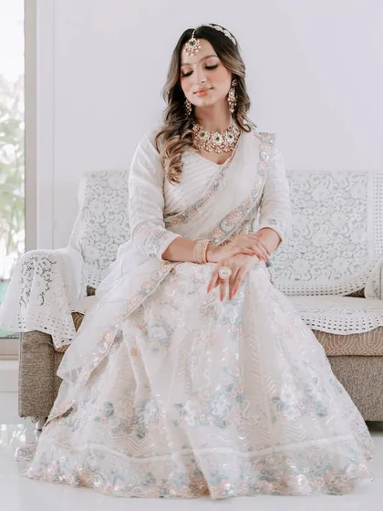 A full-length portrait of elegance. This model is seated, showcasing a beautiful white saree paired with a kundan and green stone necklace and a delicate sheeshpatti for a graceful look.