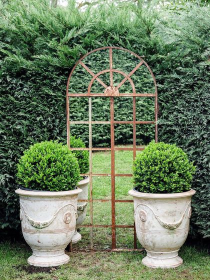 A garden mirror with a rustic frame is used to create an illusion of depth against a tall hedge. We place sculptural planters with Buxus spheres on either side to enhance the formal, symmetrical composition.