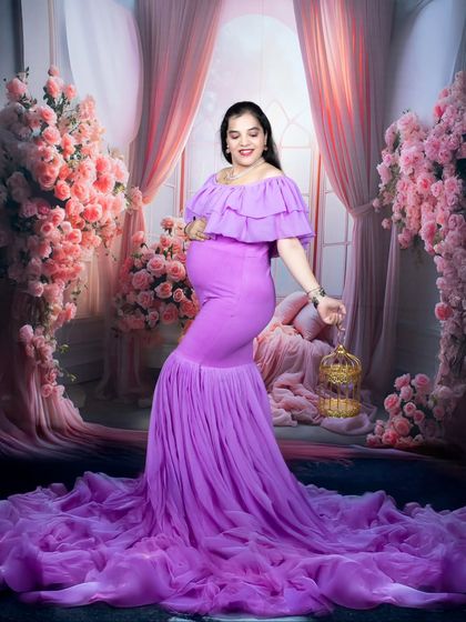 A whimsical maternity portrait of a woman in a vibrant purple mermaid-style gown, standing in a fairytale-like floral setting.