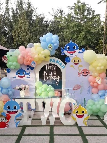 A fun and colorful Baby Shark theme for an outdoor second birthday party. The setup includes character cutouts of the whole shark family, 'TWO' marquee letters, and a pastel balloon arrangement.