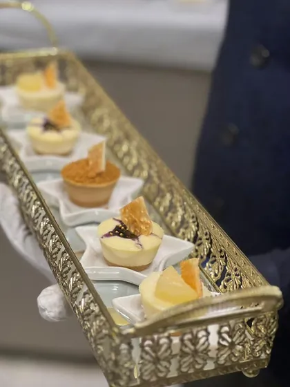A tray of assorted mini cheesecakes being served. The ornate gold tray and the server's white gloves add to the feeling of a premium, luxury service.