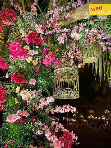 A close-up of the floral arch, showing a mix of bright pink flowers, lush greenery, and a decorative golden birdcage hanging as an accent.